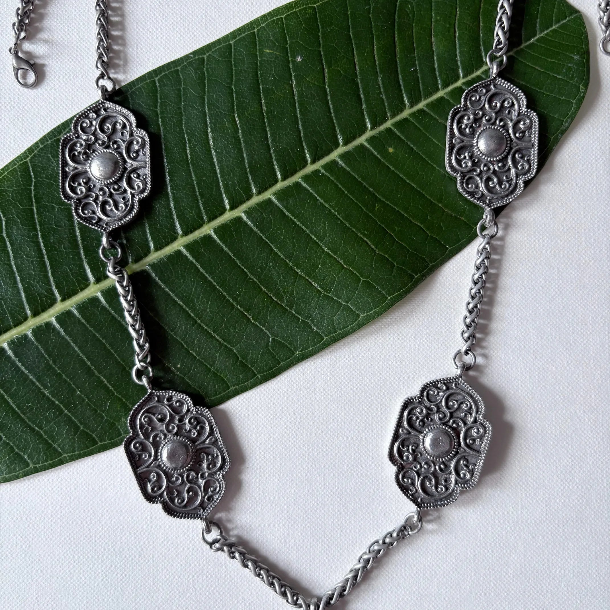 Chandrapushpa oxidised brass tribal necklace with multiple engraved motifs and dangling charms, displayed on a green leaf – Complimento artisan jewelry.