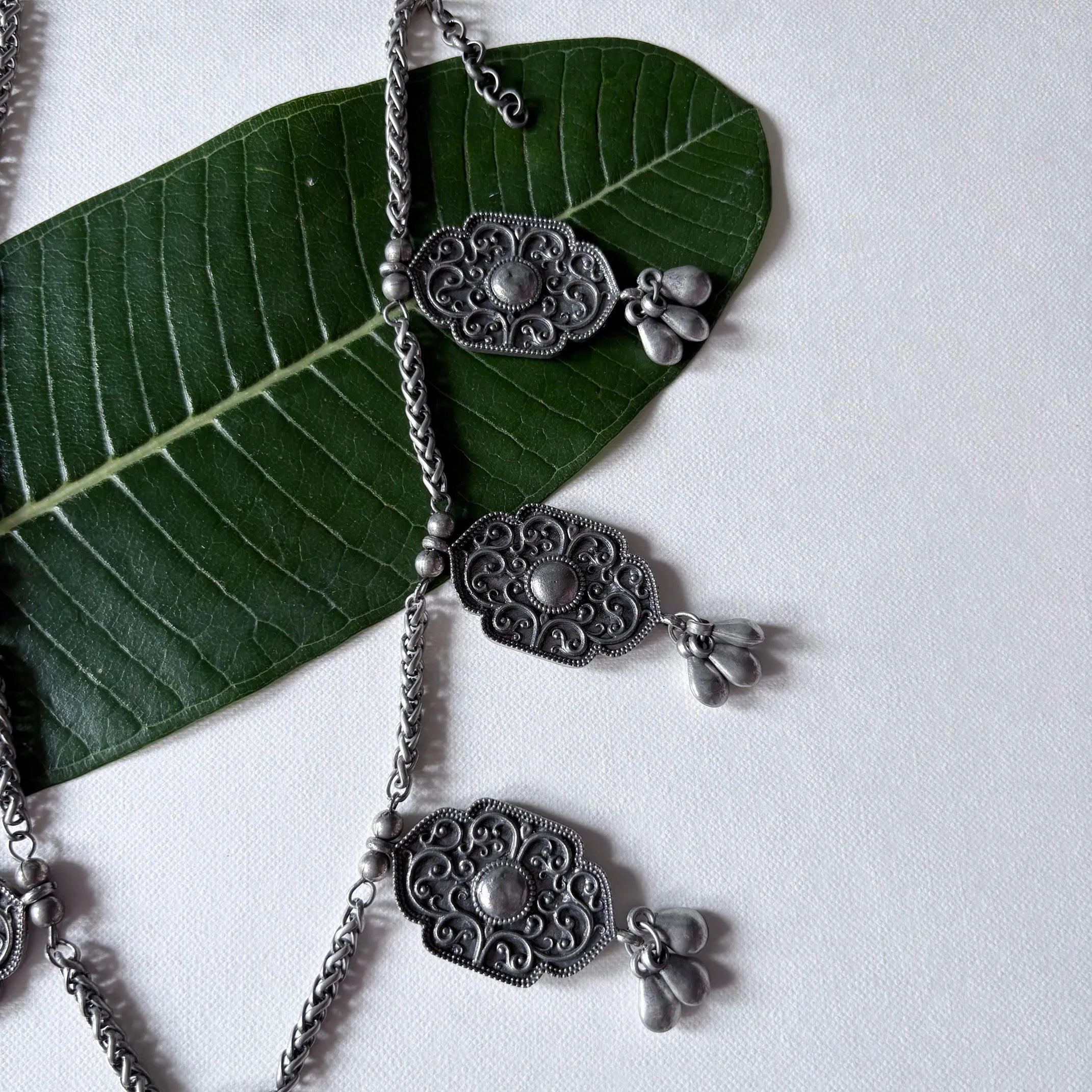 Close-up of Chandrapushpa oxidised brass necklace with engraved tribal motifs and dangling charms, displayed on a green leaf – Complimento handmade jewelry.
