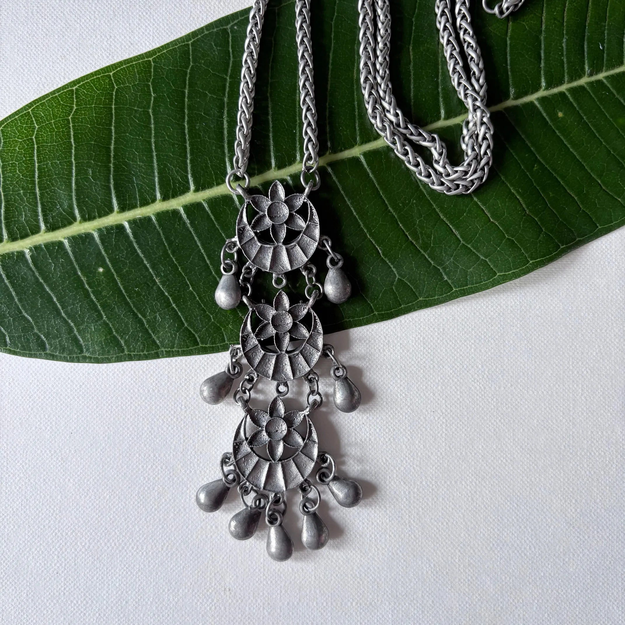 Chandrapushpa oxidised brass pendant necklace with engraved floral motif and antique finish, displayed on a green leaf – Complimento handcrafted accessory.