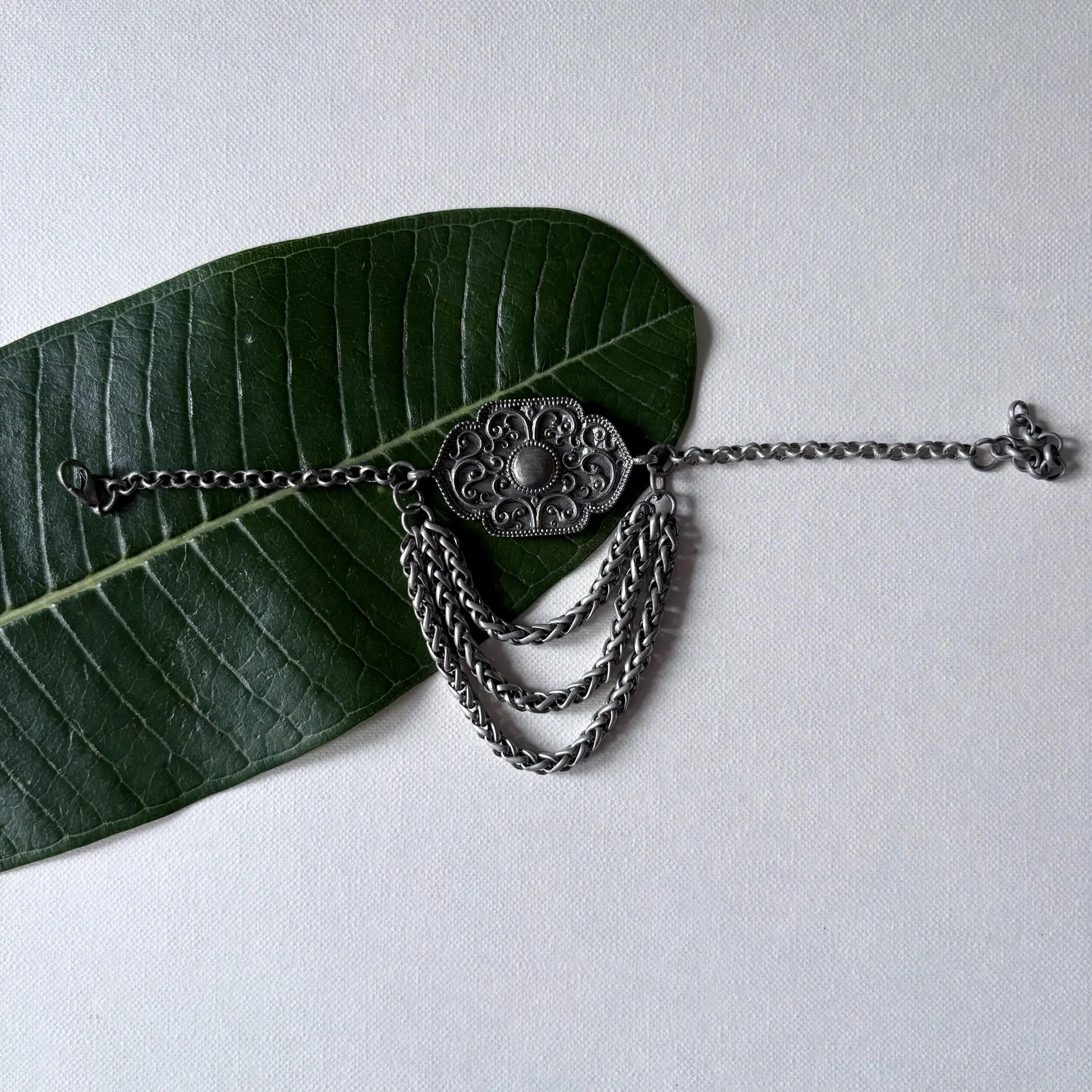 Rekha oxidised brass tribal chain bracelet with engraved motif and cascading chains, displayed on a green leaf – Complimento handcrafted jewelry.
